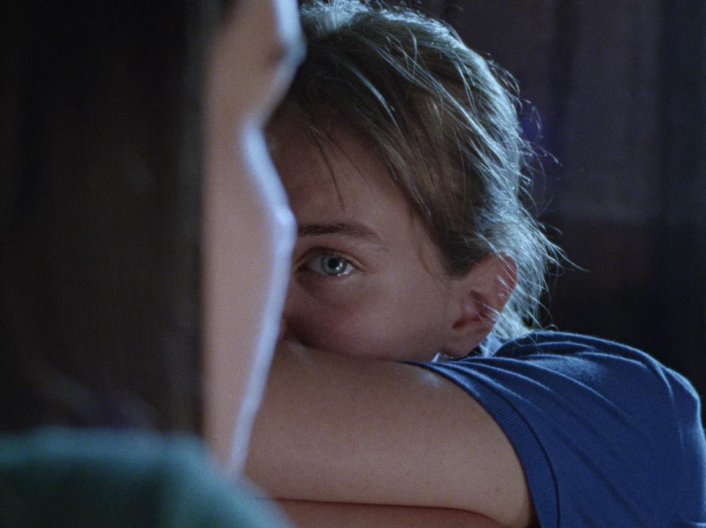 A young girl buries her face in her arms and looks at a woman in the foreground.