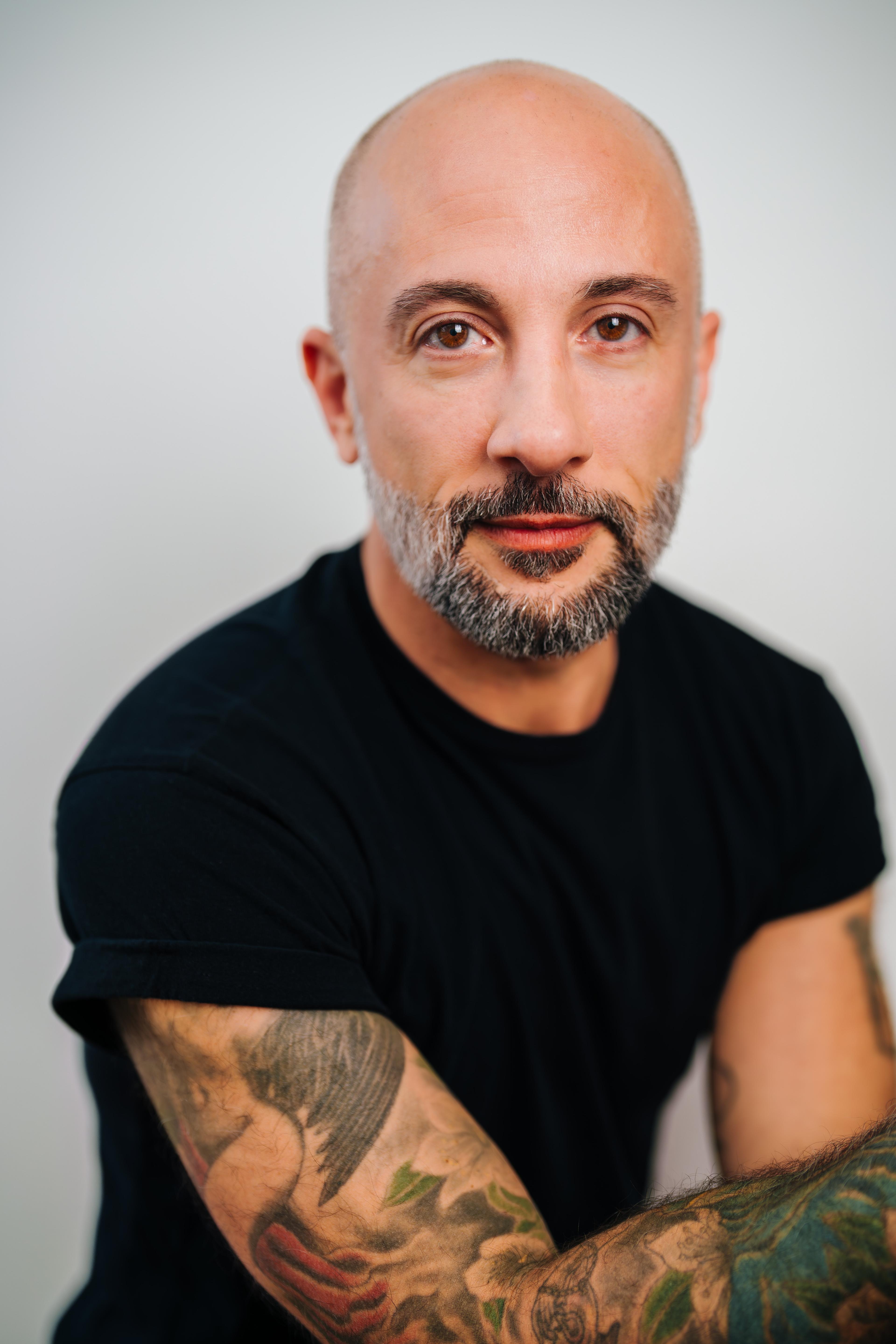 An olive-skinned, bald person with a short salt-and-pepper beard, wears a black T-shirt. They face the camera with a calm, direct expression, arms folded, showing tattooed forearms against a neutral light background.