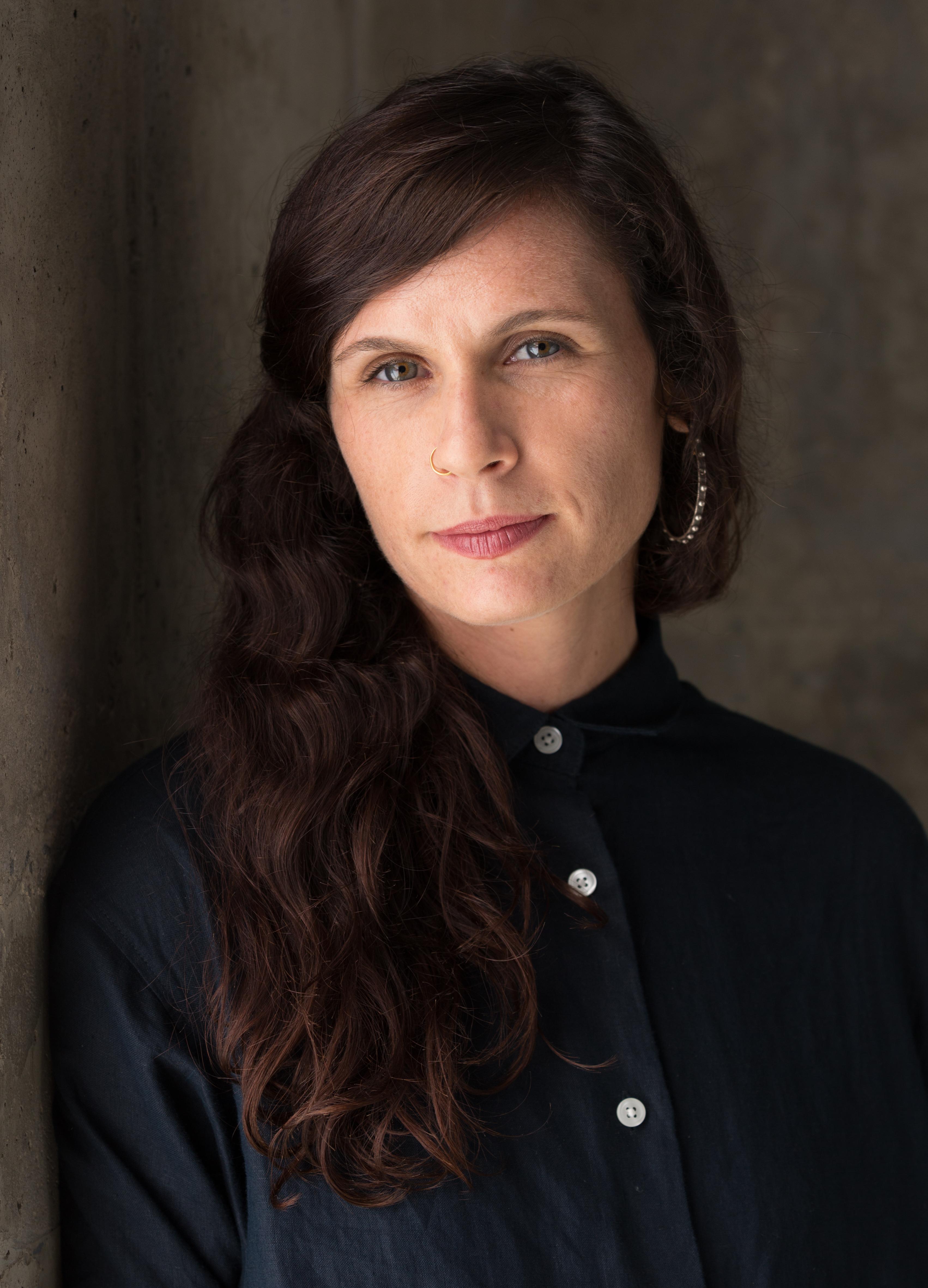 Headshot of a woman with long wavy dark hair, gold nose ring, wearing a dark button-up shirt against a concrete wall.