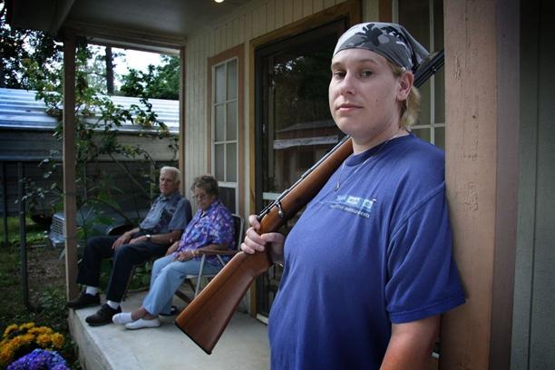 A woman wearing a bandana holds a shotgun over her shoulder and looks into the camera. In the background, an elderly couple sits on their porch.