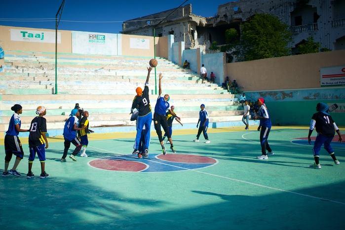 A group of children and teenagers play basketball on a colorful court. Two players reach up for the ball suspended in air.
