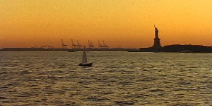 The Statue of Liberty overlooks the Hudson River at sunset. A sailboat floats by.