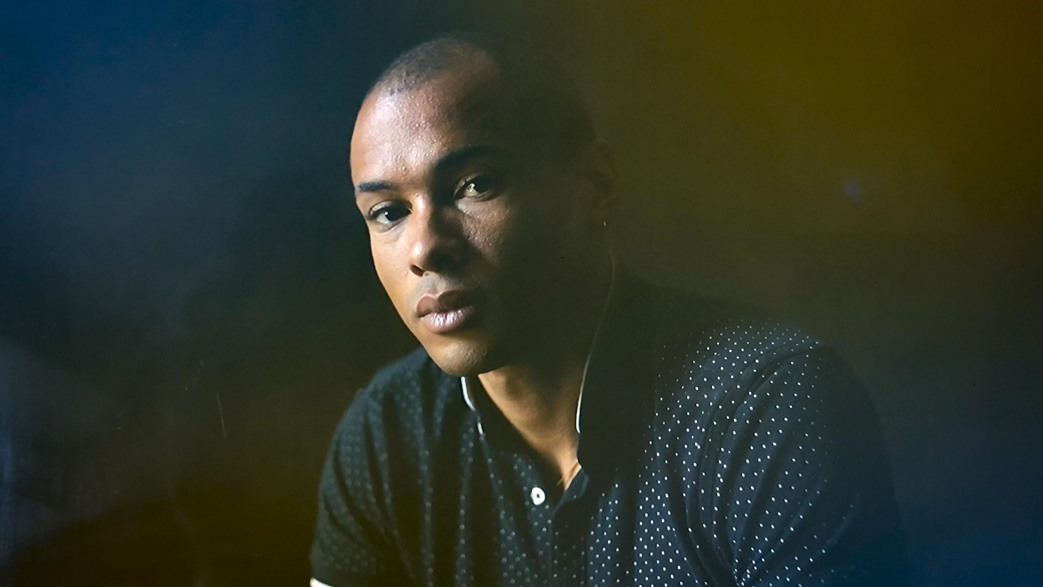 A portrait of a man in front of a dark backdrop with a yellow and blue filter.