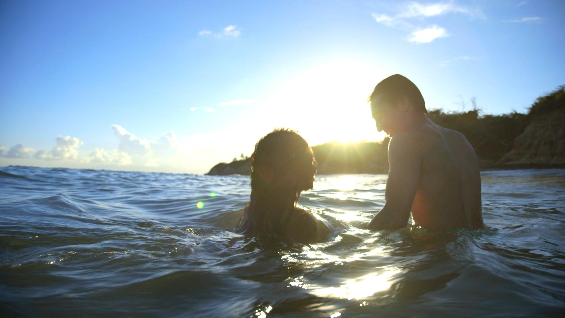 A man and women swim in the ocean together as the sun shines brightly in the background