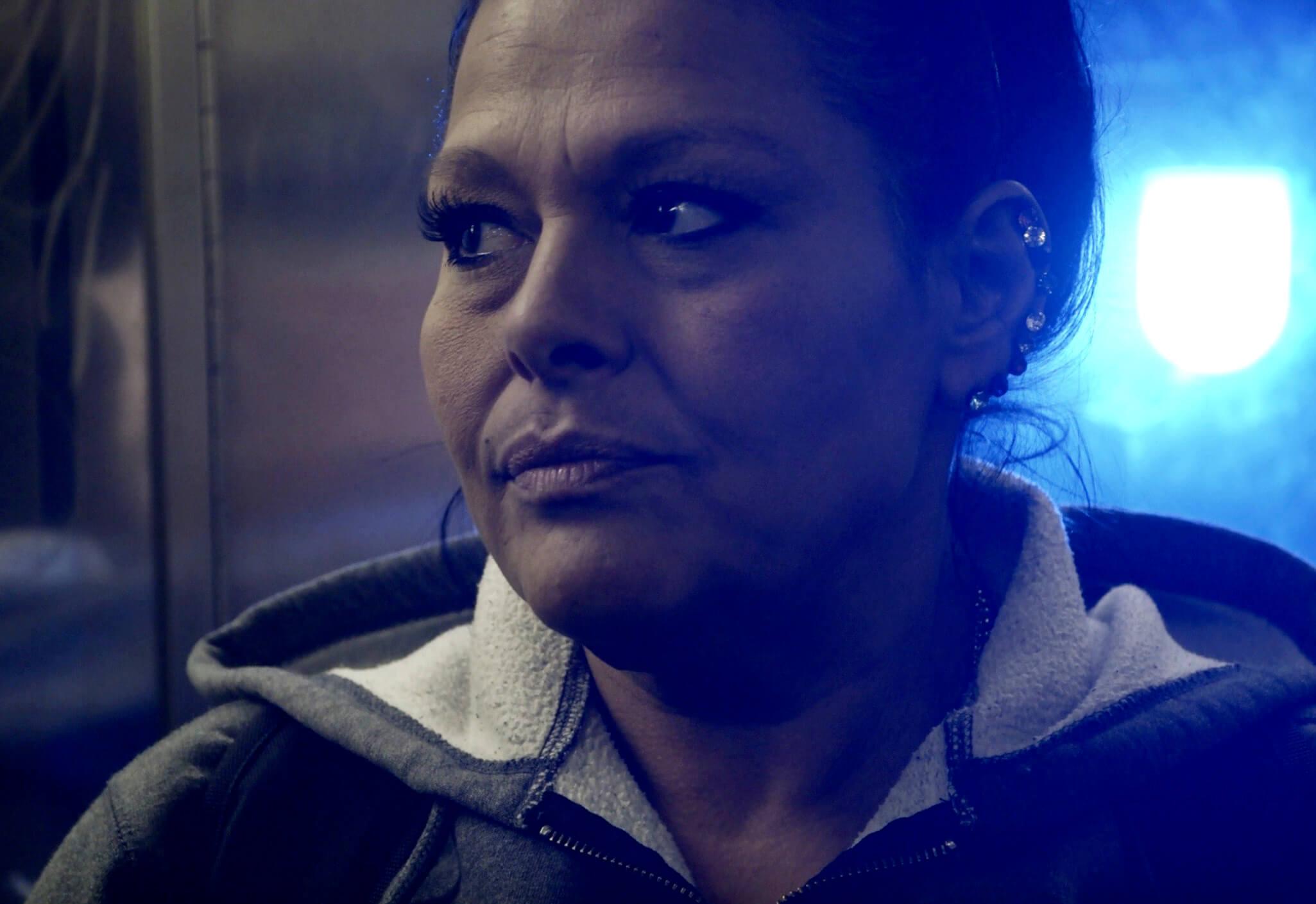 Still from And So I Stayed. A woman looks off to the side as she is bathed in blue light from the backround.