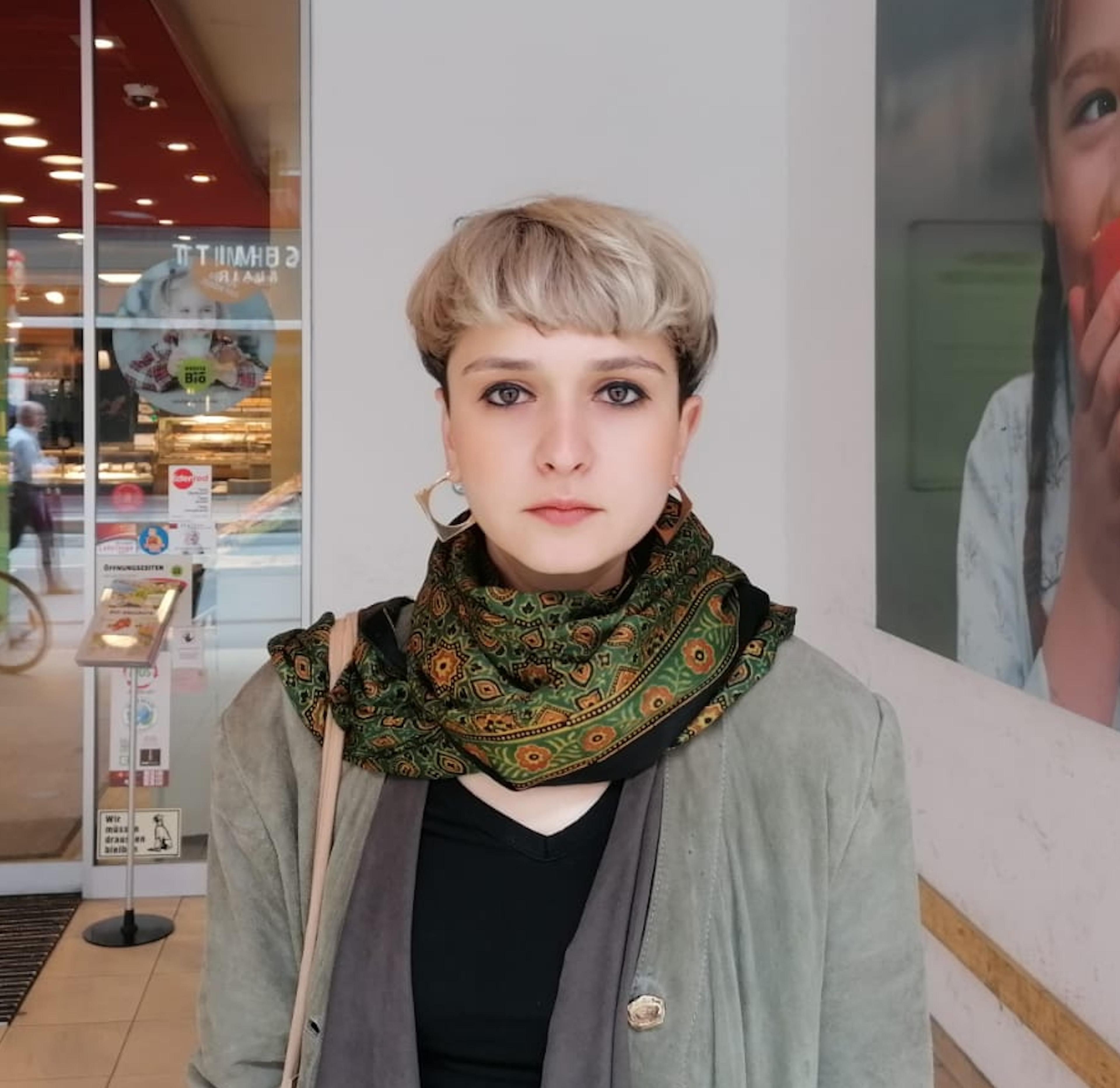Cristina Hanes — Portrait of filmmaker Cristina Hanes (Romania), NoCut Film Collective. A woman with a short blonde pixie cut, wearing a green patterned scarf and grey jacket, standing outside a shopfront.