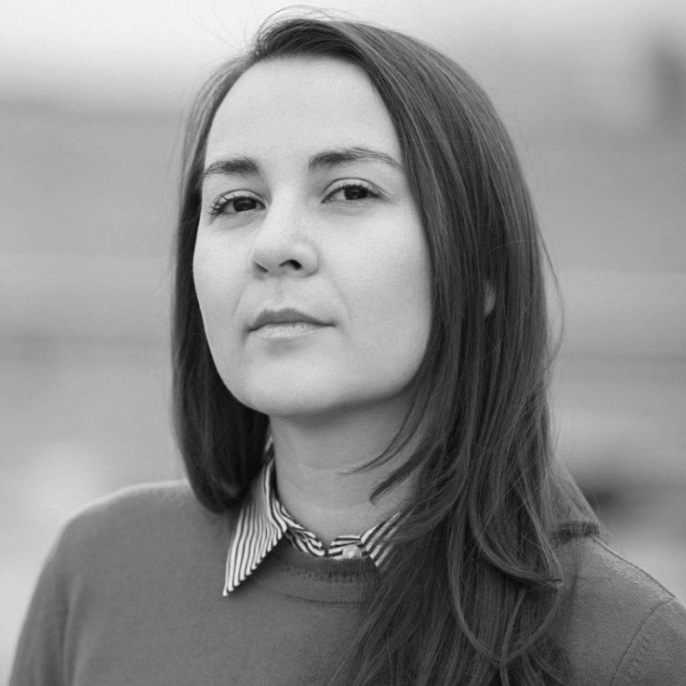 A black and white portrait of filmmaker Brooke Pepion Swaney. A young woman with shoulder length dark hair looks intently at the camera. She wears a collared shirt and a crew neck sweater over it.