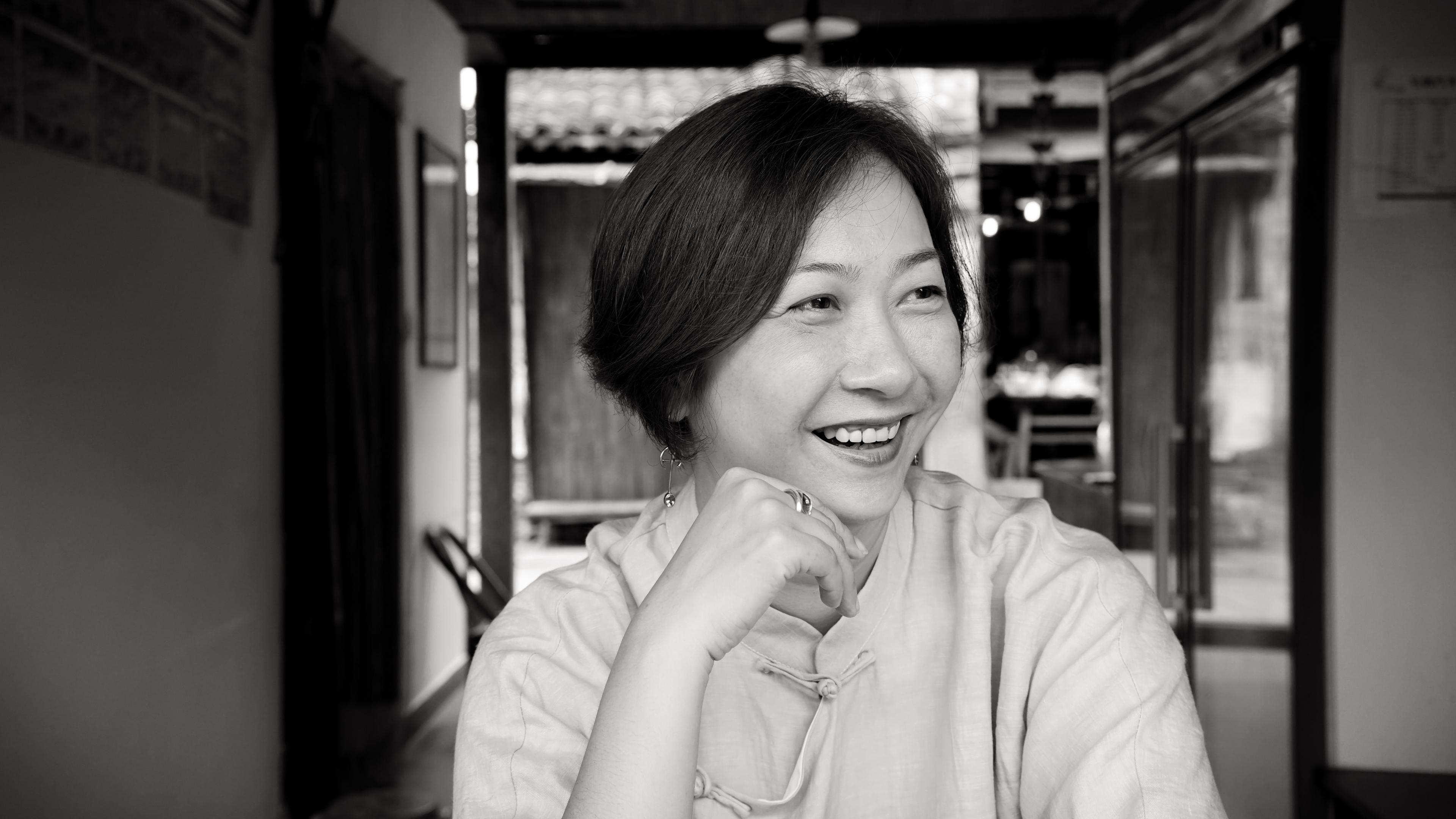 Zhao Qing looking to her side and smiling. She has short dark hair, and is wearing a light-colored blouse. Black and white portrait, with out of focus background.