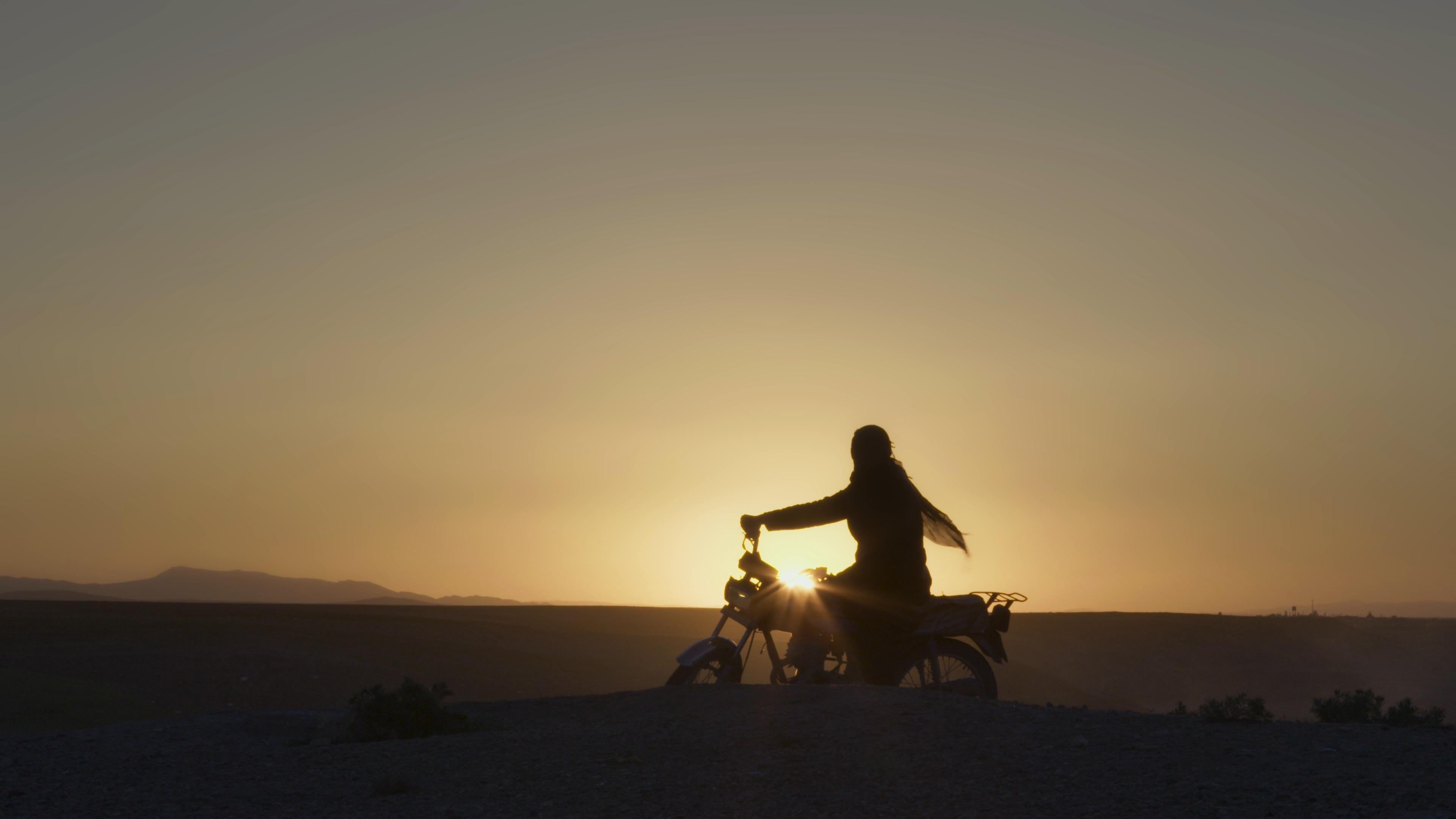 In the Iranian desert, a woman sits on a motorcycle and overlooks the landscape. She is backlit by the setting sun.