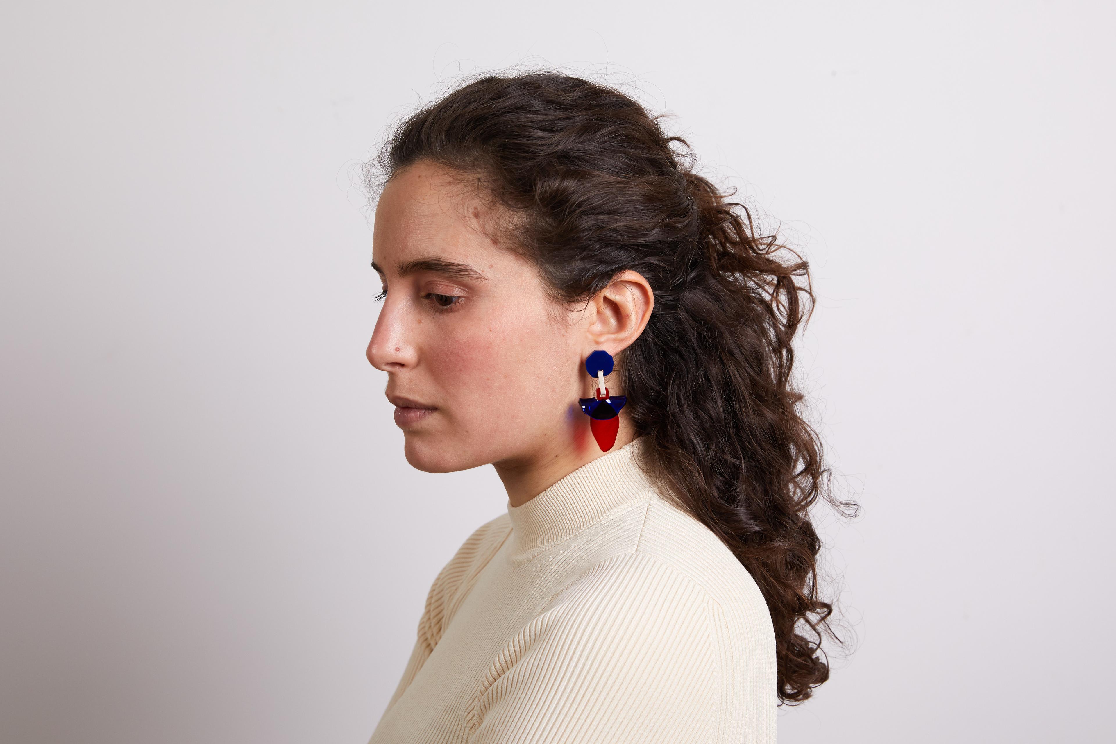 Headshot of a woman's profile as she is turned away from the camera. She has curly brown hair, and is wearing red and blue earrings, and an off-white long sleeve shirt.