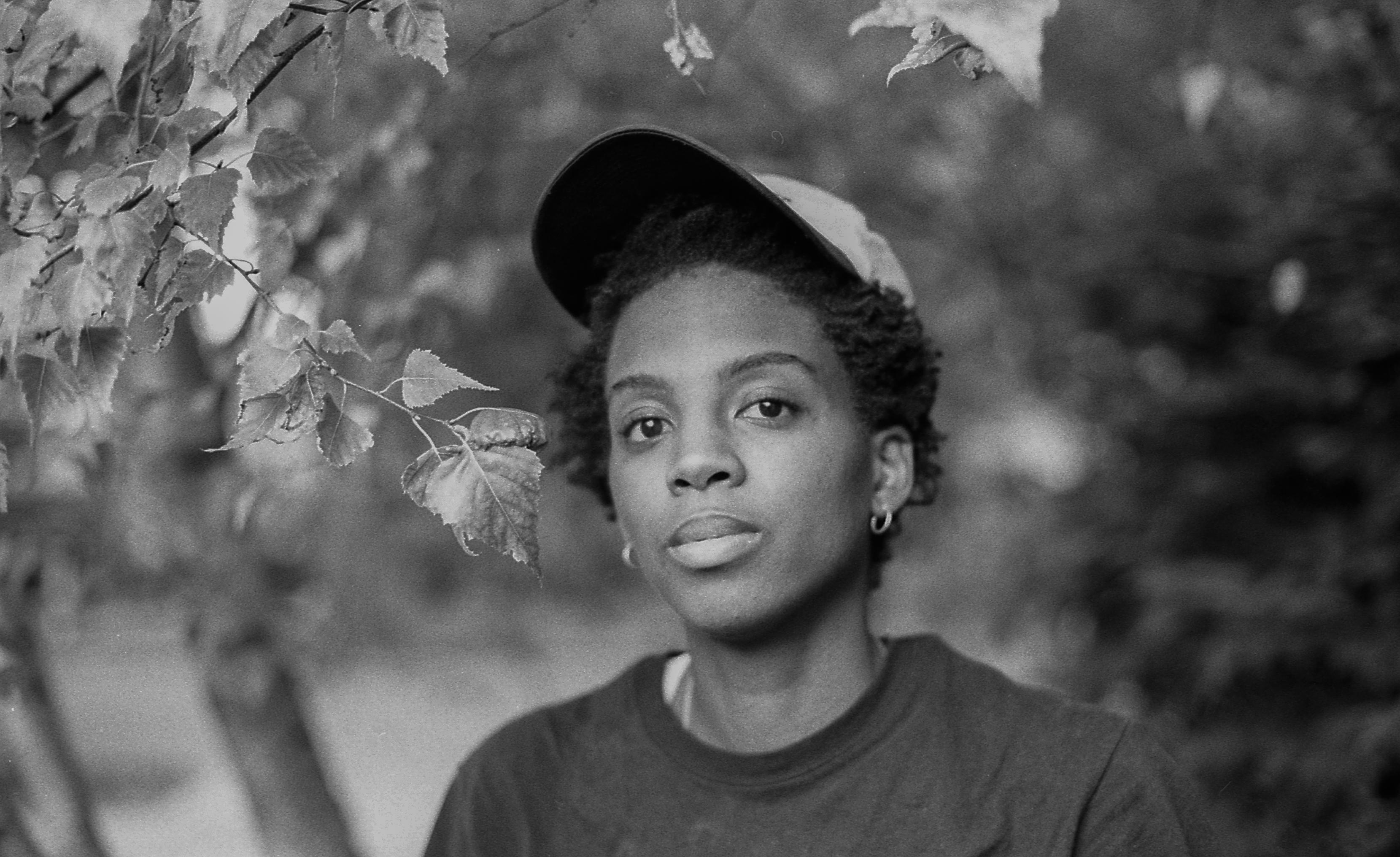 Cai Thomas looks directly at the camera. She has short hair and wears a cap. Portrait in black and white.