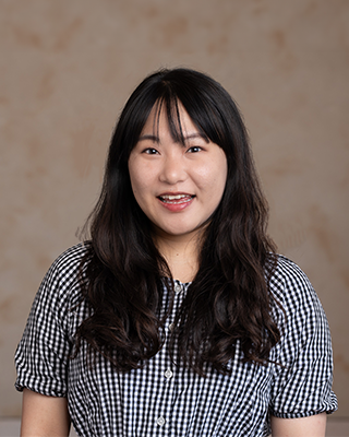 A Korean woman filmmaker smiles warmly at the camera in a studio portrait. She has long dark hair styled in loose waves with bangs and wears a black-and-white gingham-patterned dress. The softly textured beige background creates a simple and professional setting.