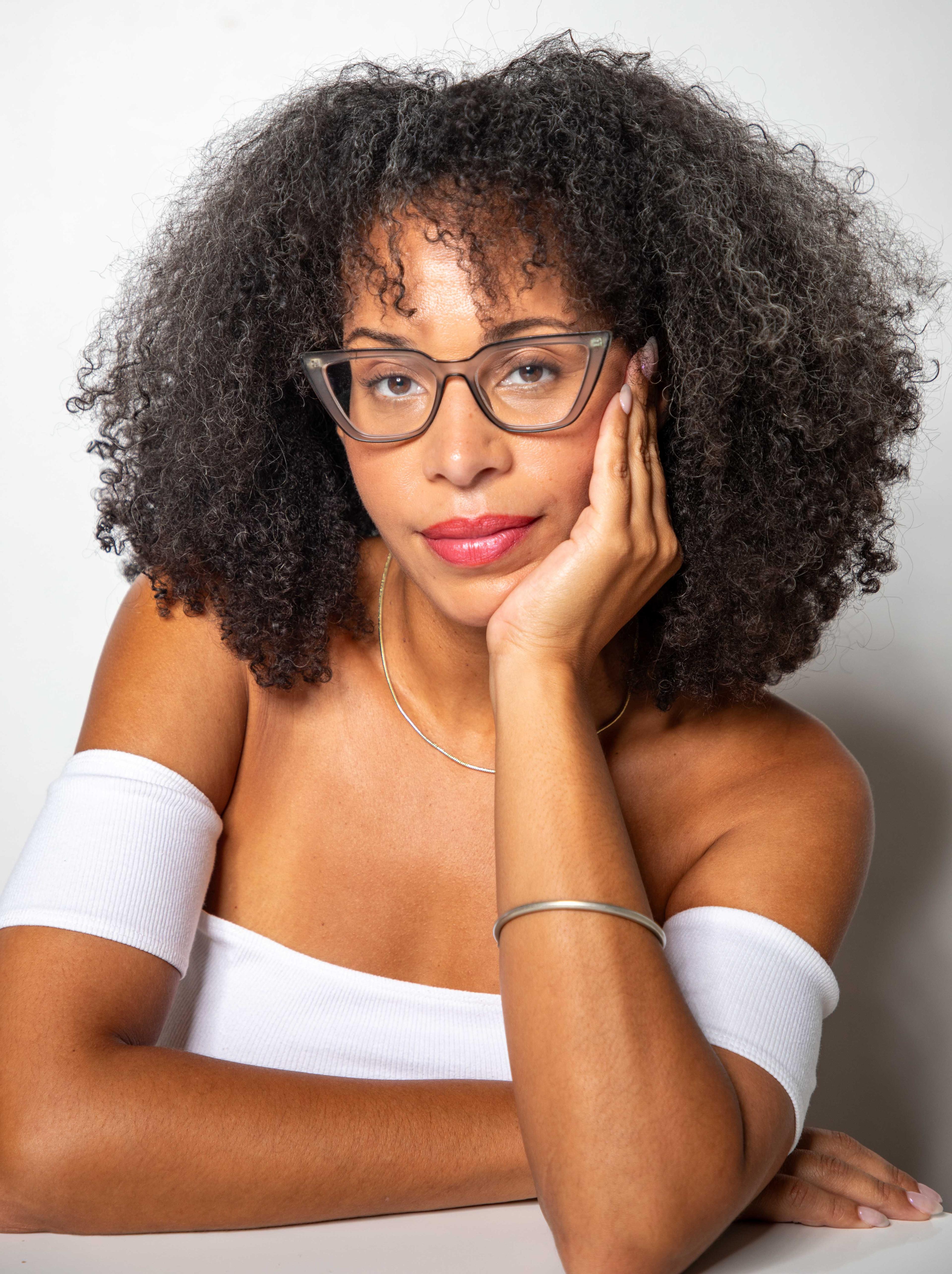 Loira Limbal is looking directly at the camera. She is wearing a white dress and glasses, with her head resting on her hand.