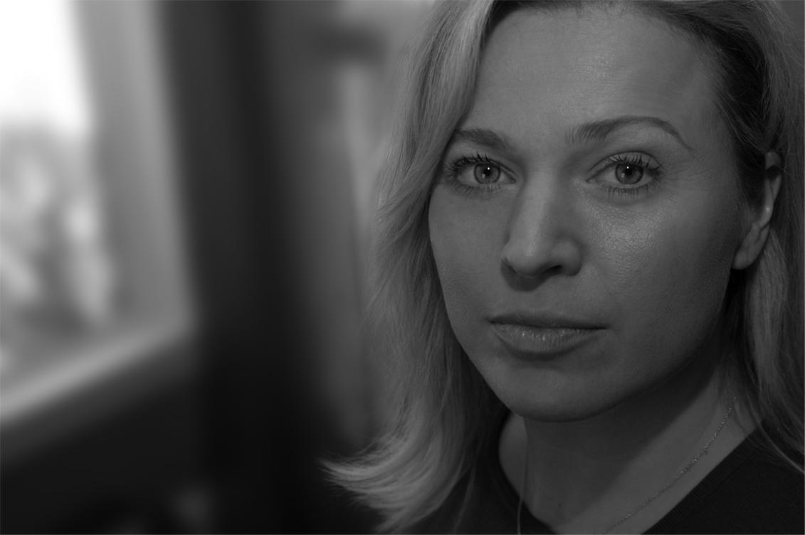 A black and white photo of a woman next to a window looking into the camera.