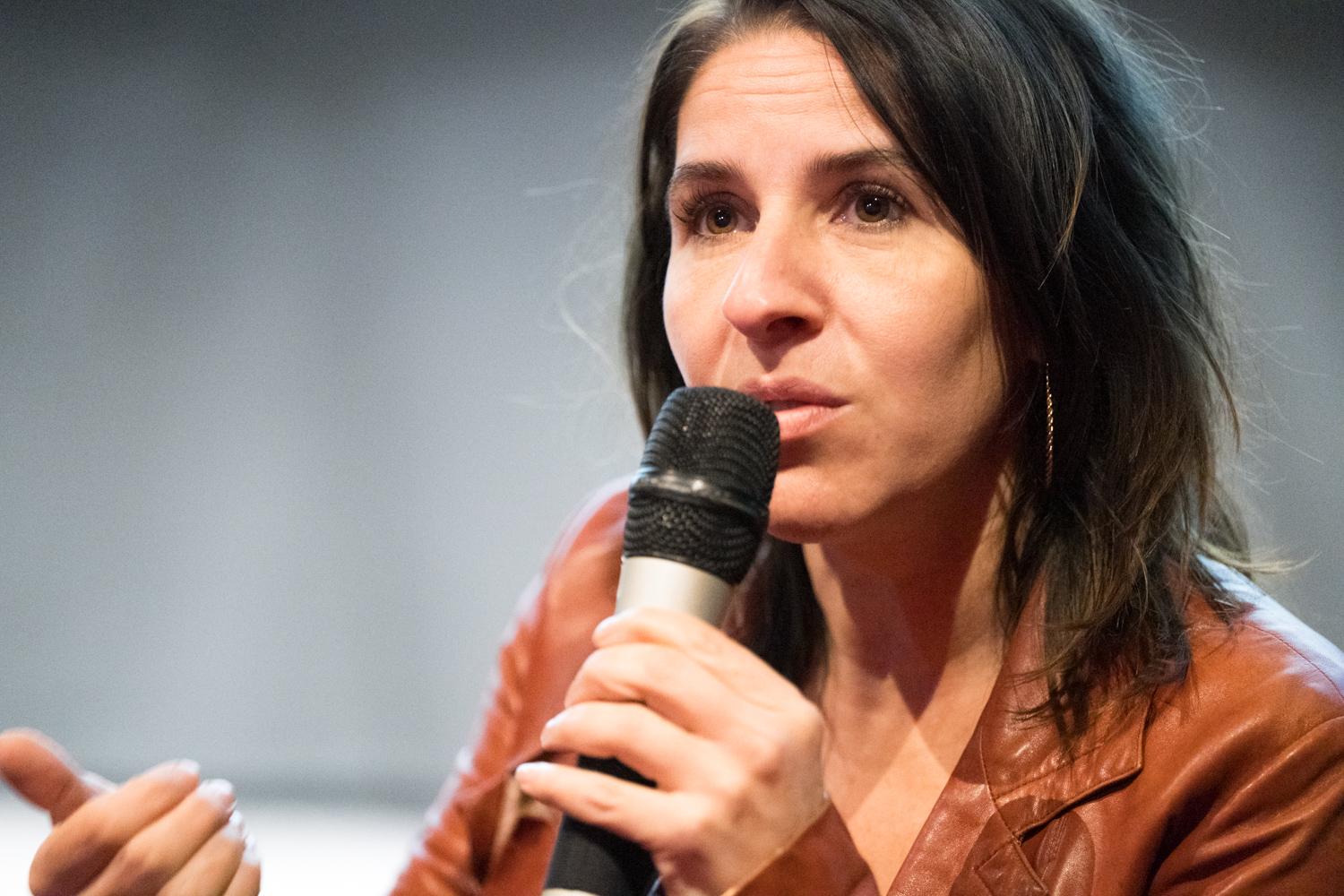 Close up portrait of woman with short brown hair wearing a light brown leather jacket. She is holding a microphone and speaking into it as she motions with her hand, seemingly looking at an audience (not pictured).