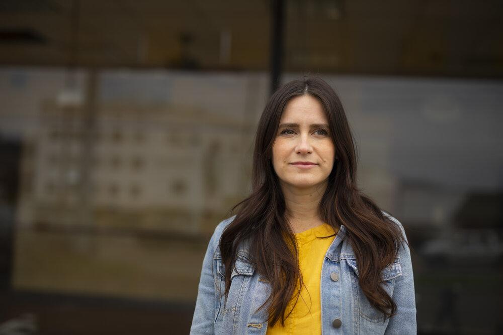 A portrait of a woman wearing a bright yellow shirt and a denim jacket in front of a window.