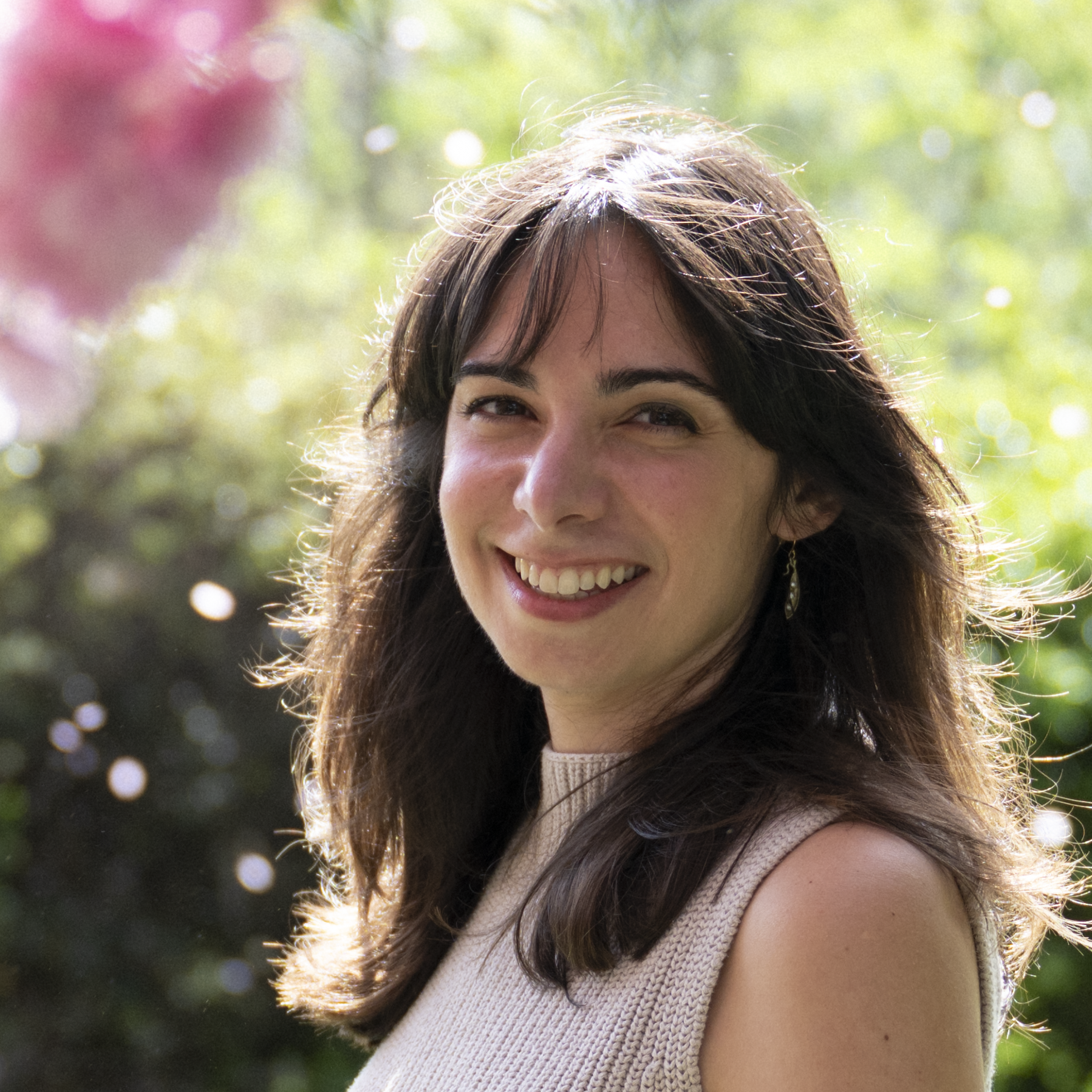 A woman with long dark brown hair and wispy bangs smiles warmly at the camera over her shoulder. She wears a sleeveless cream knit turtleneck top and small gold drop earrings. The photo is taken outdoors with a softly blurred background of green foliage and a pink flower, with natural sunlight creating a bright, airy feel.