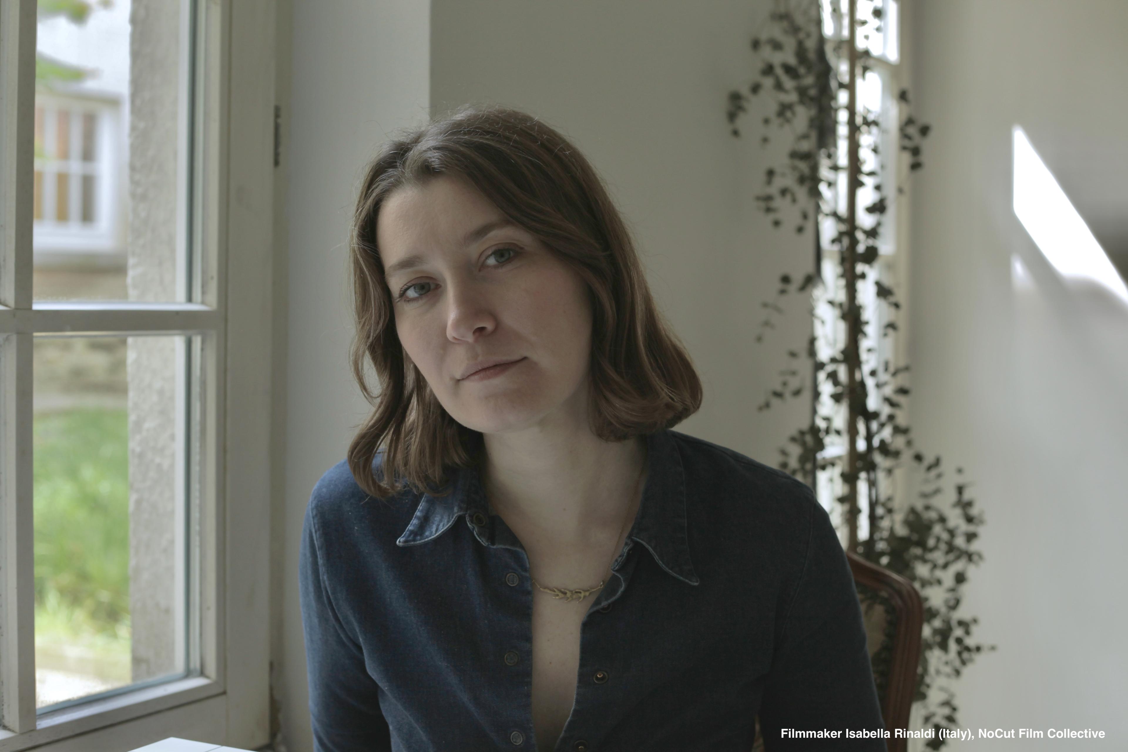 Isabella Rinaldi — Portrait of filmmaker Isabella Rinaldi (Italy), NoCut Film Collective. A woman with shoulder-length brown hair, wearing a blue denim shirt, seated by a window in soft natural light.