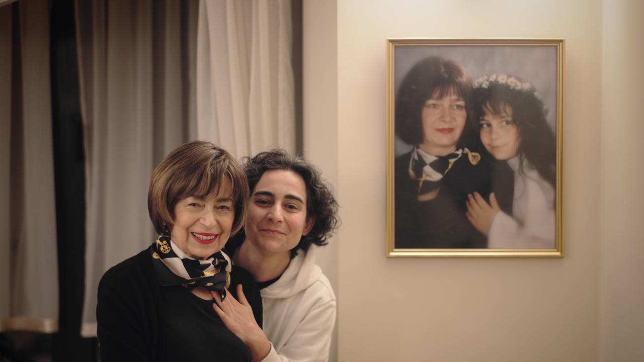 A woman and her adult son stand closely together indoors, smiling warmly at the camera. The older woman on the left has short brown hair and wears a black outfit with a patterned scarf. A person beside her has short curly hair and rests her hand affectionately on the older woman's shoulder. On the wall to their right hangs a framed portrait of what appears to be the same two individuals years earlier—an adult woman with short dark hair and a little girl wearing a white dress and a flower crown, posed together.