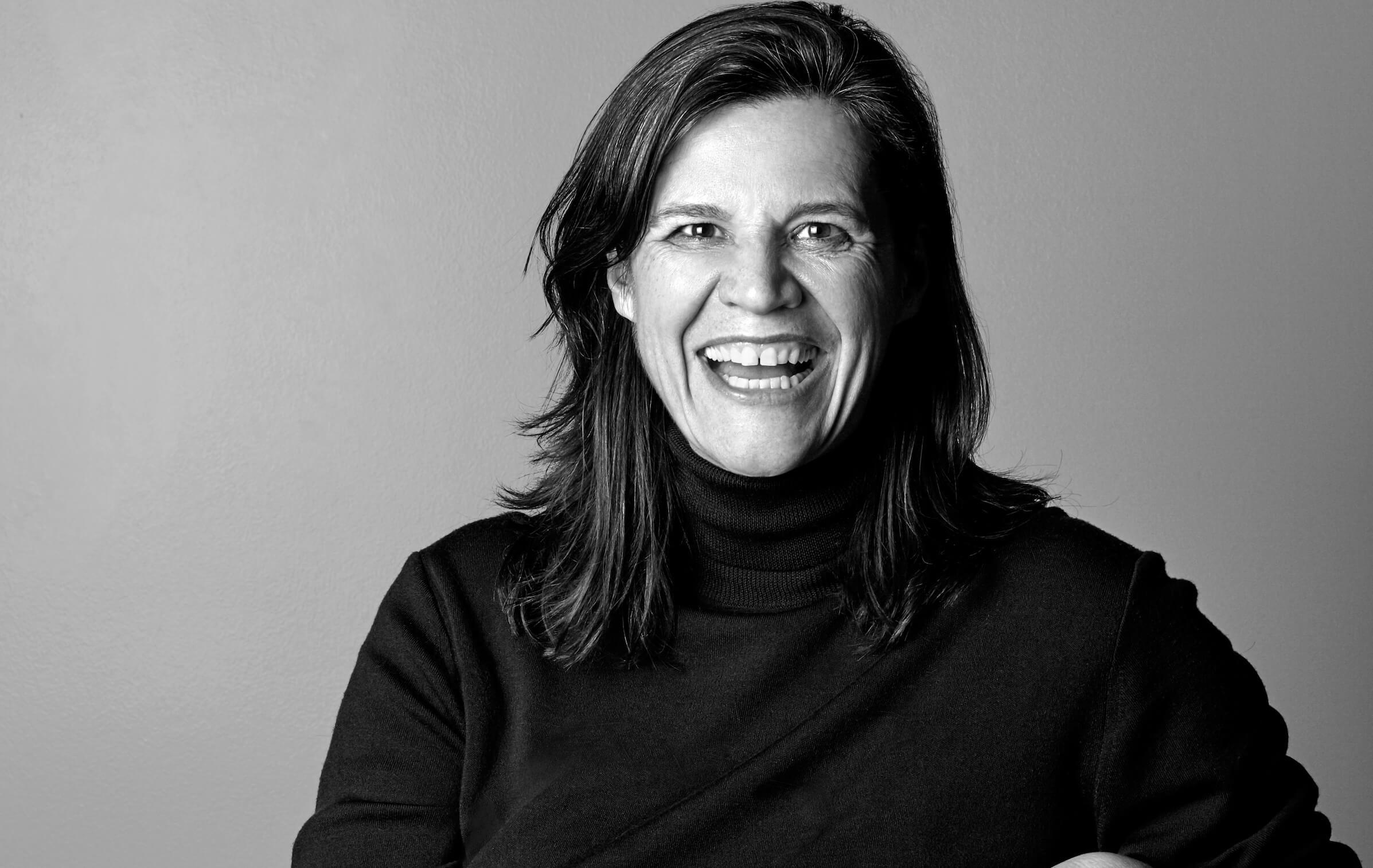 Kirsten Johnson sitting in a chair, wearing metallic pants and a turtleneck shirt. She has medium hair and is widely smiling at the camera. Black and white portrait.