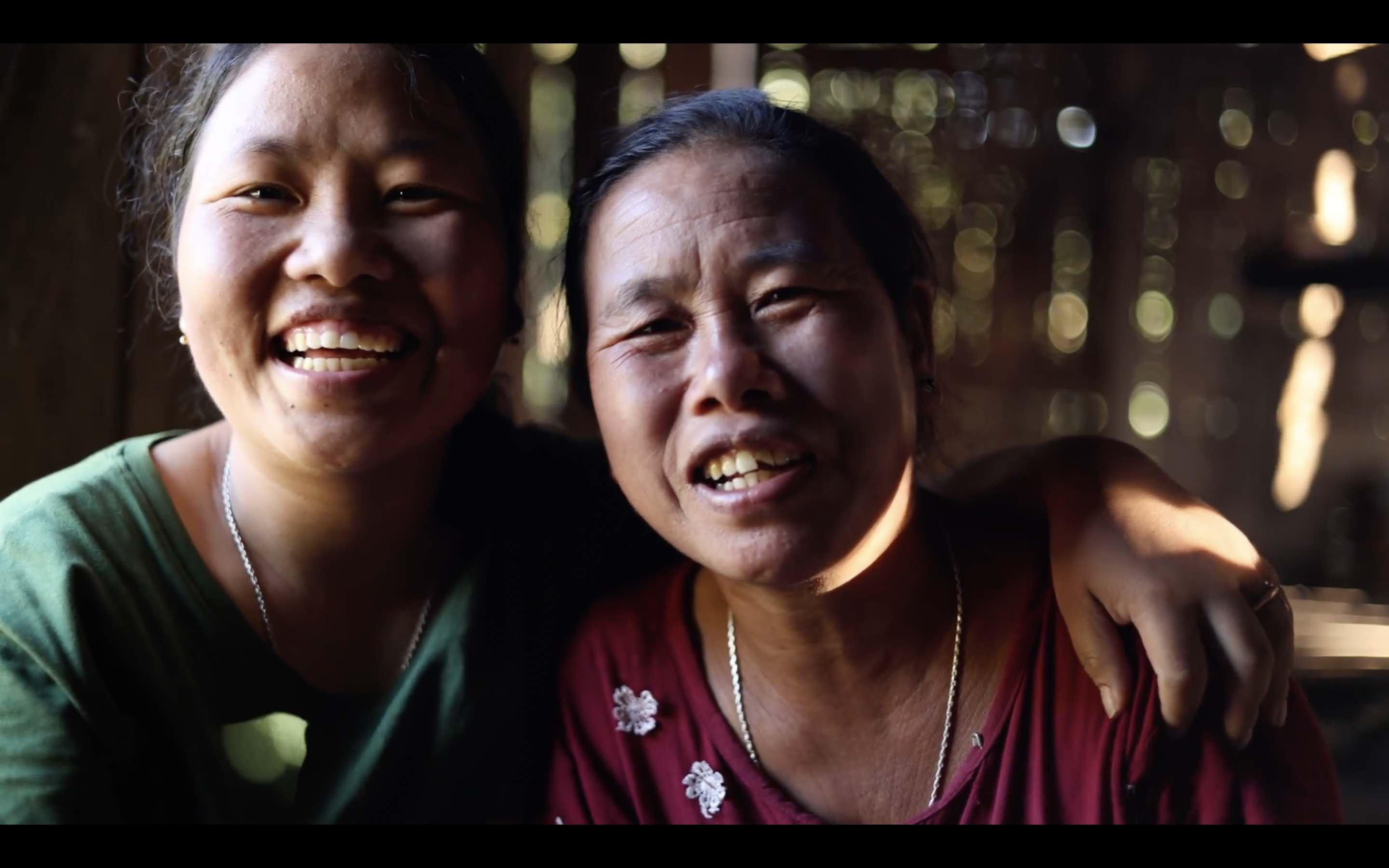 A young Miju Mishmi woman and her mother are sitting in a thatch house, looking directly into the camera and smiling