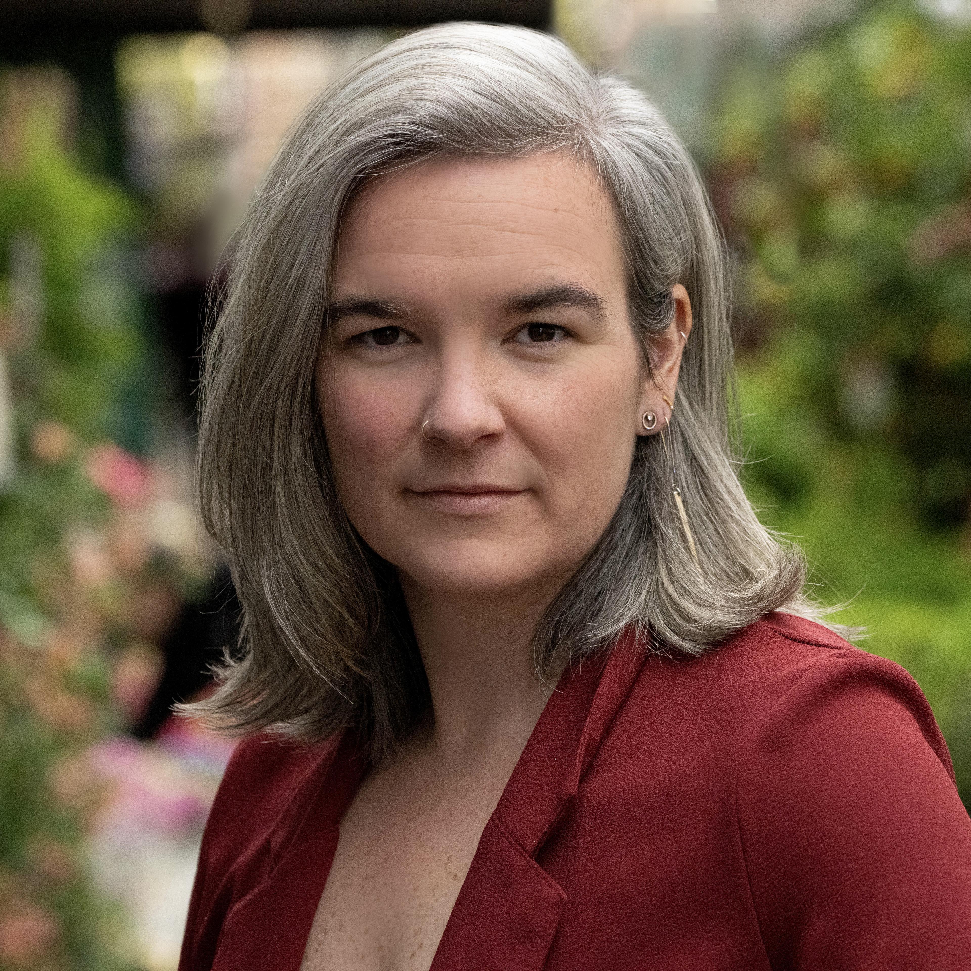 Professional headshot of a woman with shoulder-length gray hair, wearing a deep red blazer, photographed outdoors against a softly blurred green garden background.
