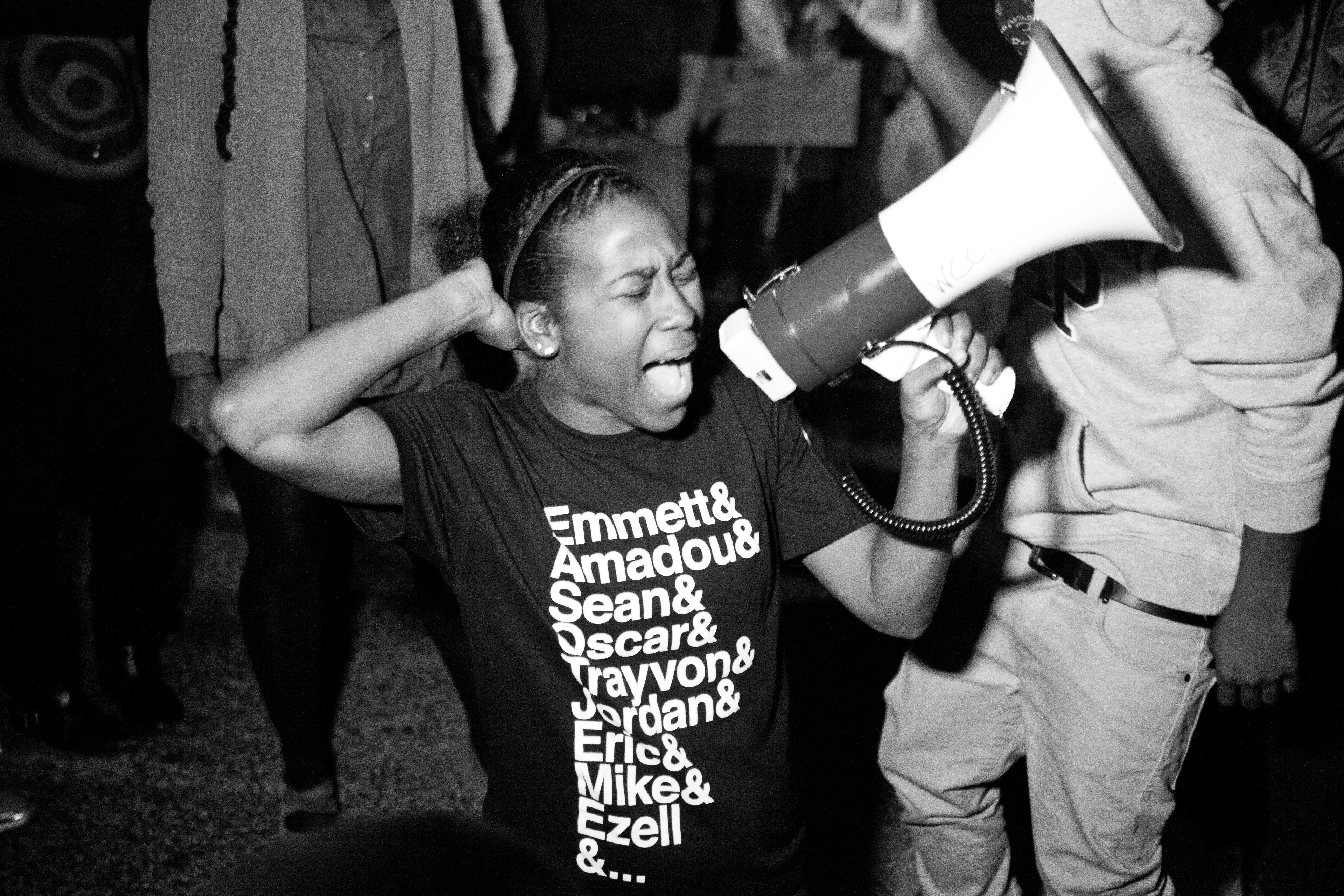 Film still from Whose Streets?, a 2016 (Egg)celerator Lab grantee by filmmakers Sabaah Jordan and Damon Davis. A young African American woman shouts into a loudspeaker. She wears a t-shirt with the names of many African American men who have been killed by police forces.