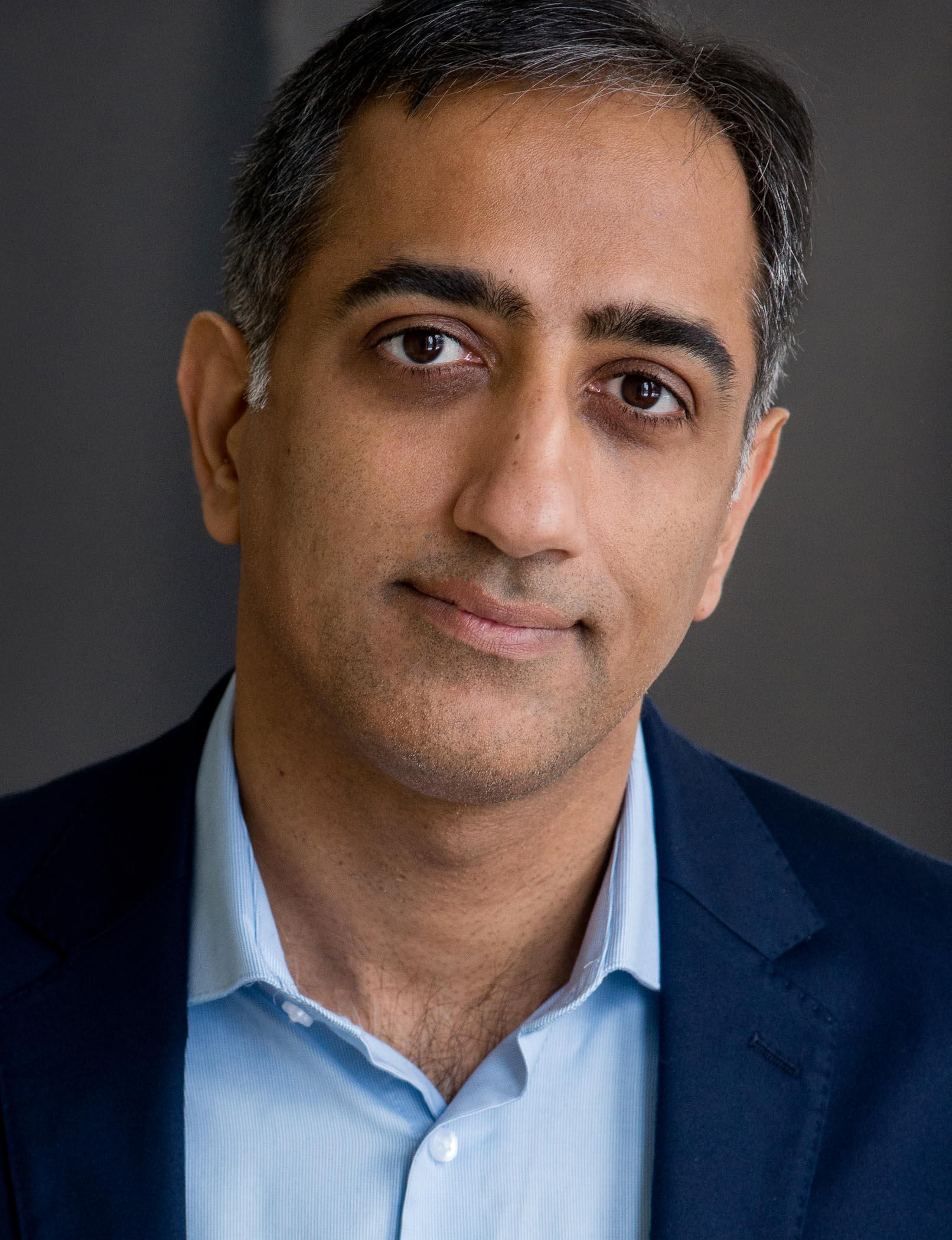 A headshot of Amar Lohana, who is a middle-aged South Asian man with short salt and pepper hair. He is wearing a blue blazer and blue shirt.
