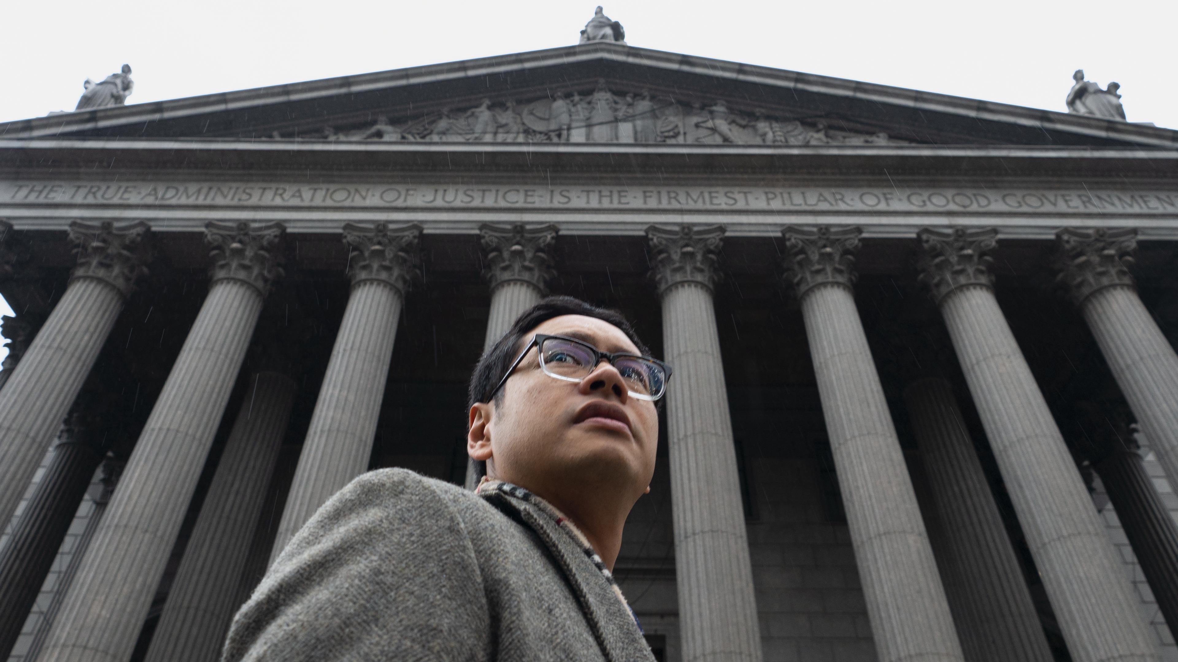 A low-angle image of a man wearing glasses and a formal outfit standing in front of a the New York State Supreme Courthouse.
