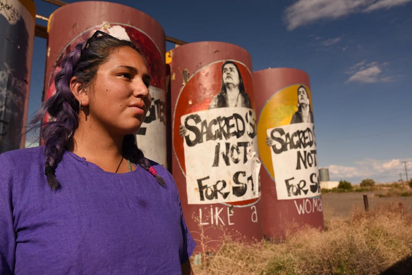 Portrait of Ivey Camille Manybeads Tso. Ivey looks away from the camera, with braided hair and a t-shirt. In the back there are columns with posters pasted that protest the sale of Native land.