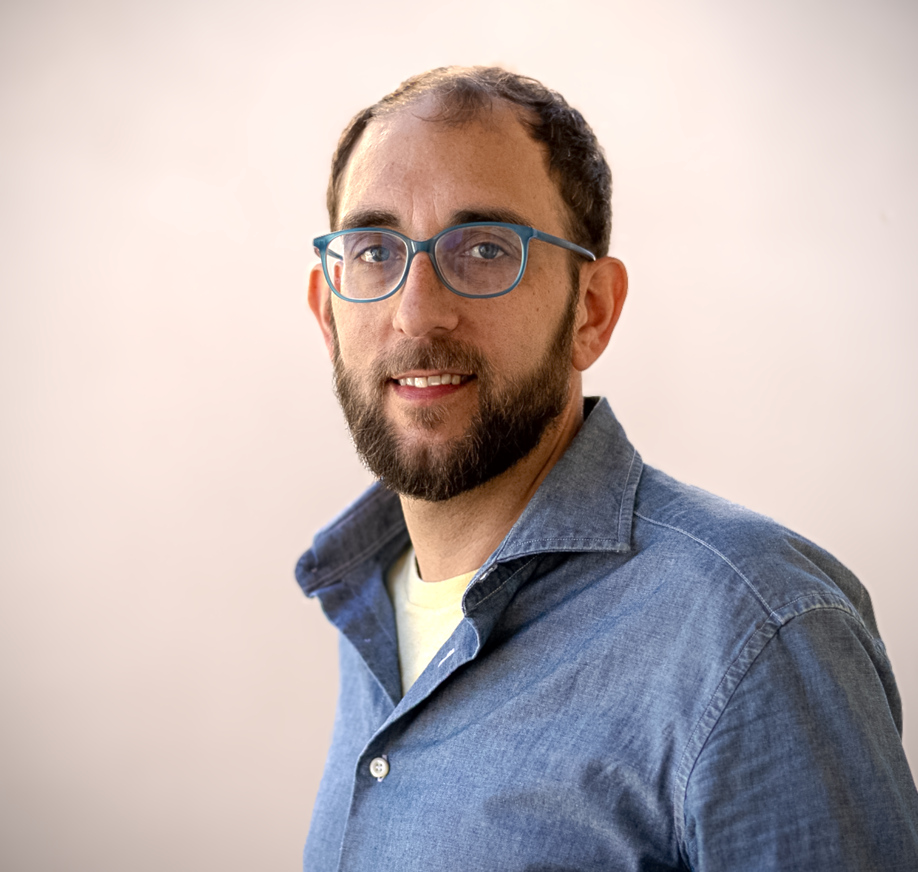 Portrait of a man with short dark hair and a trimmed beard, wearing blue-framed glasses and a blue button-up shirt. He is turned slightly to the side, facing the camera with a relaxed, friendly expression against a plain light background.