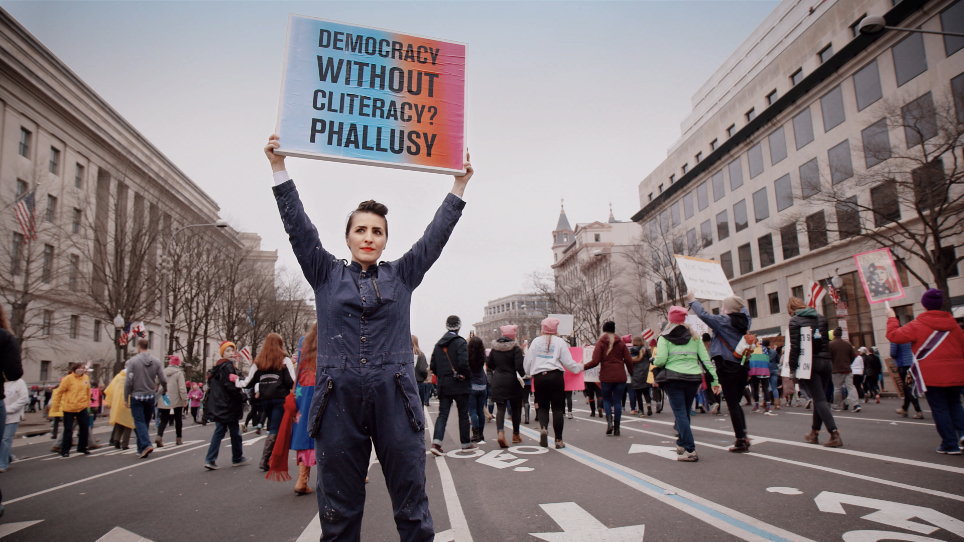 A person holds up a sign at a protest saying "Democracy without cliteracy? Phallusy"