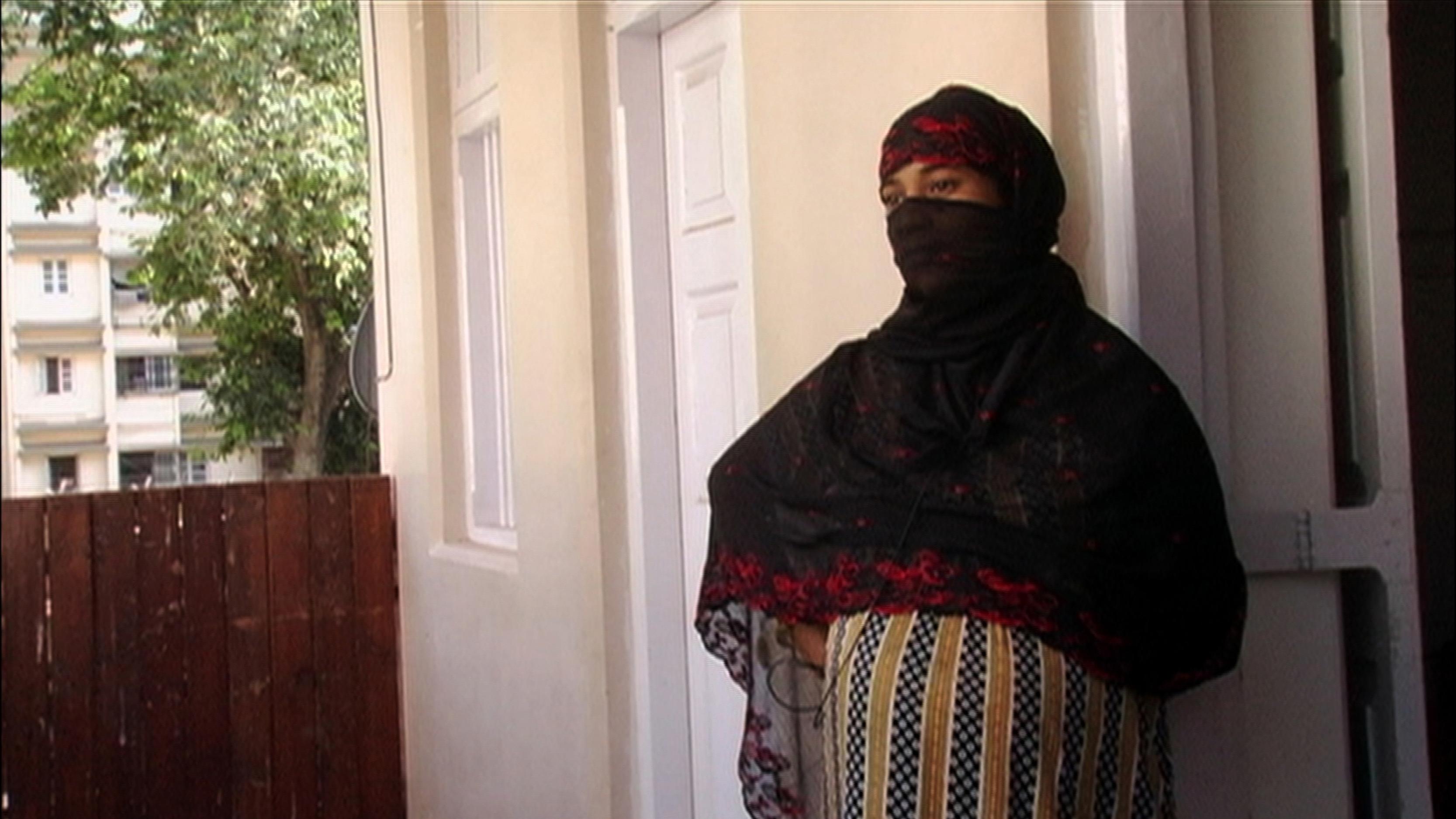 A young girl wearing a face and head covering stands on a porch.