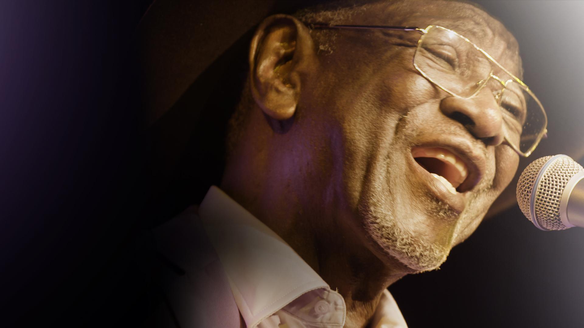 A closeup of a man wearing glasses and a hat singing into a microphone.