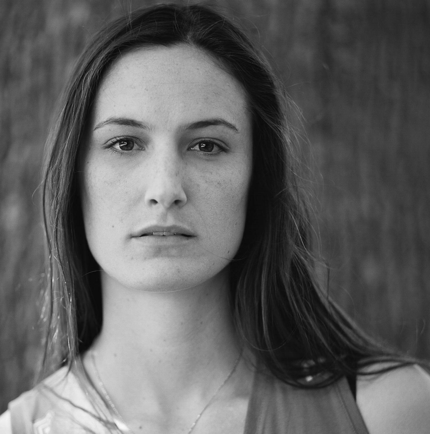 A black and white photo of a woman standing in front of a tree and looking into the camera.
