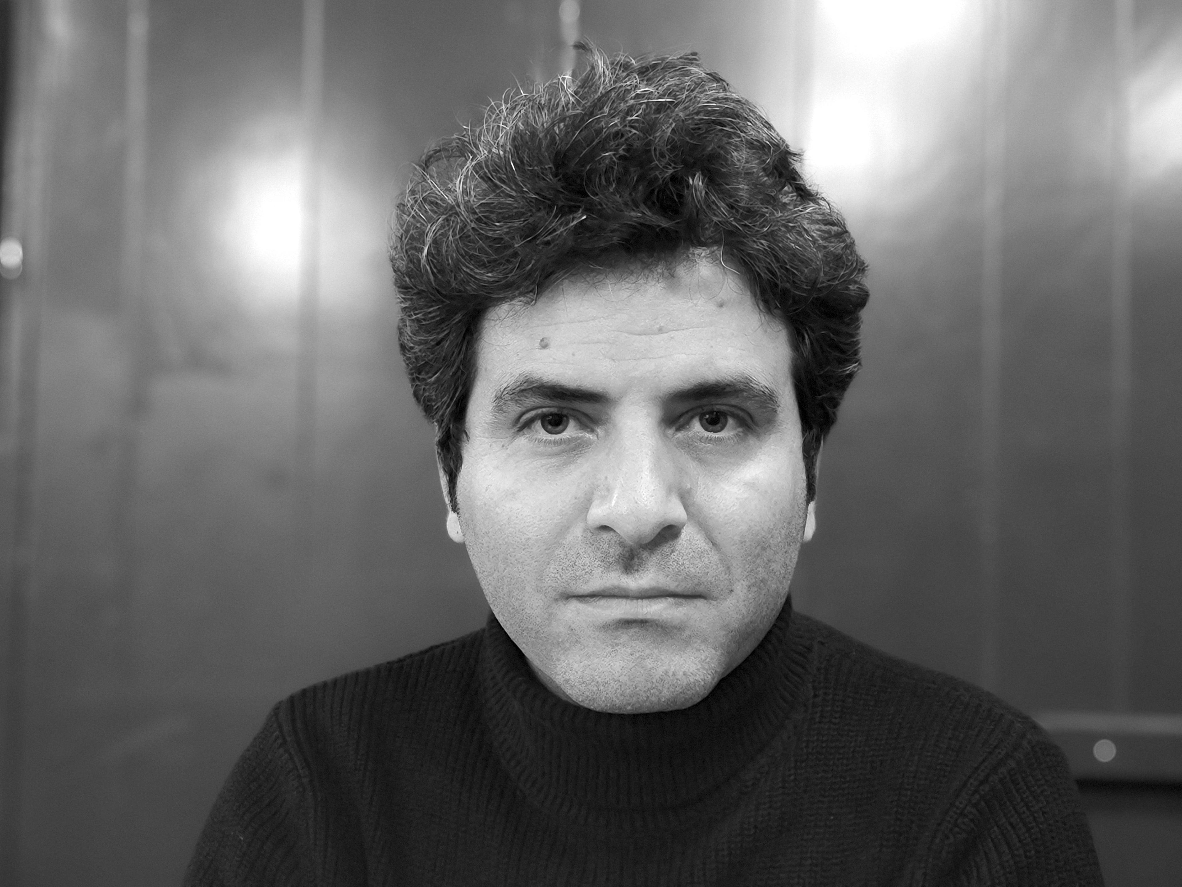 A black and white portrait of a man with bushy hair and a black turtleneck looking into the camera.