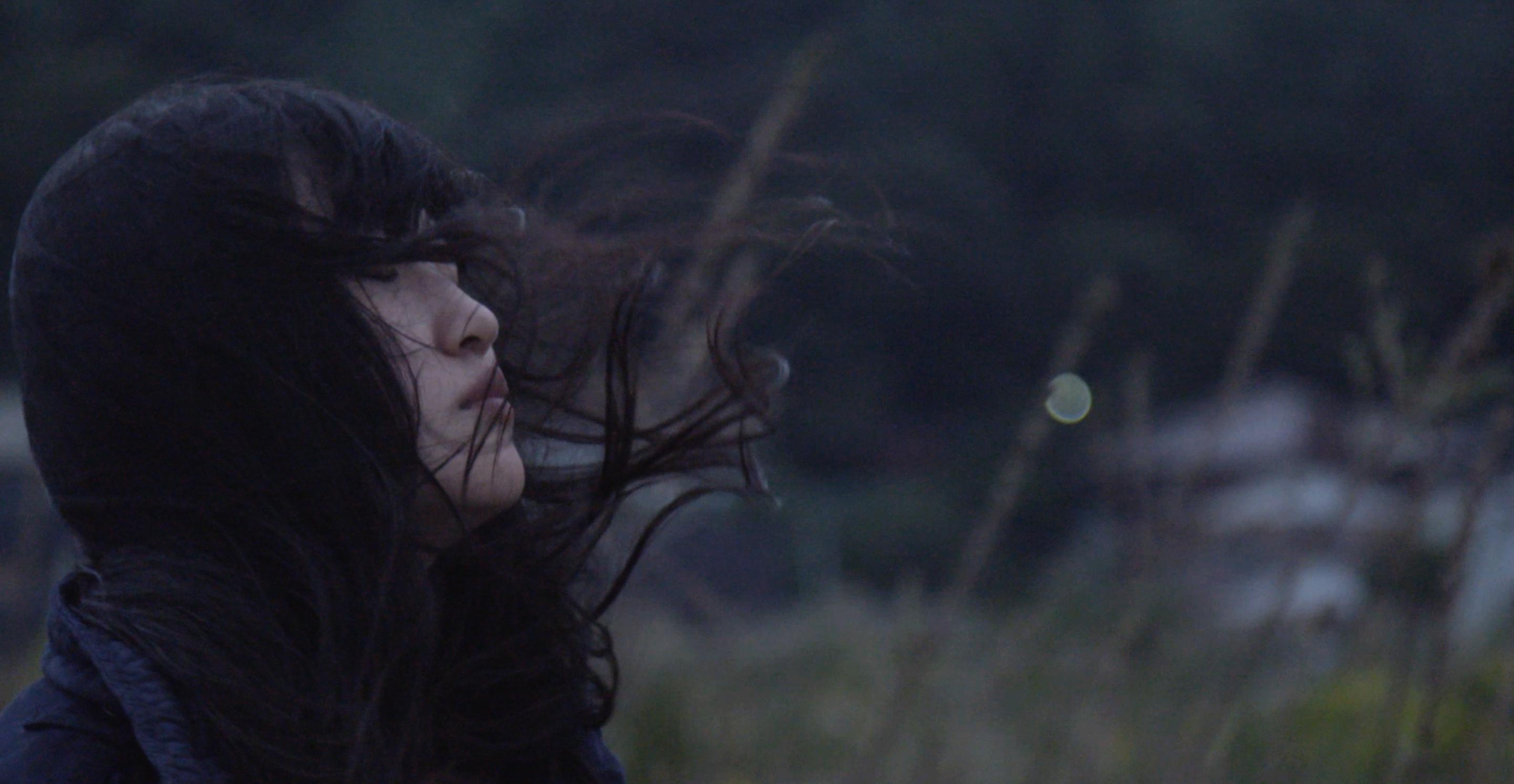 Still from All of Our Heartbeats Are Connected Through Exploding Stars. A woman closes her eyes as the wind blows her hair.