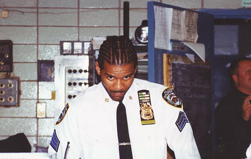 Corey Pegues, one of the highest ranking black executives in the NYPD, wearing his police uniform with many insignia and badges to indicate his seniority. his hair is braided and he looks up slightly toward the photographer while standing at a desk inside his precinct.