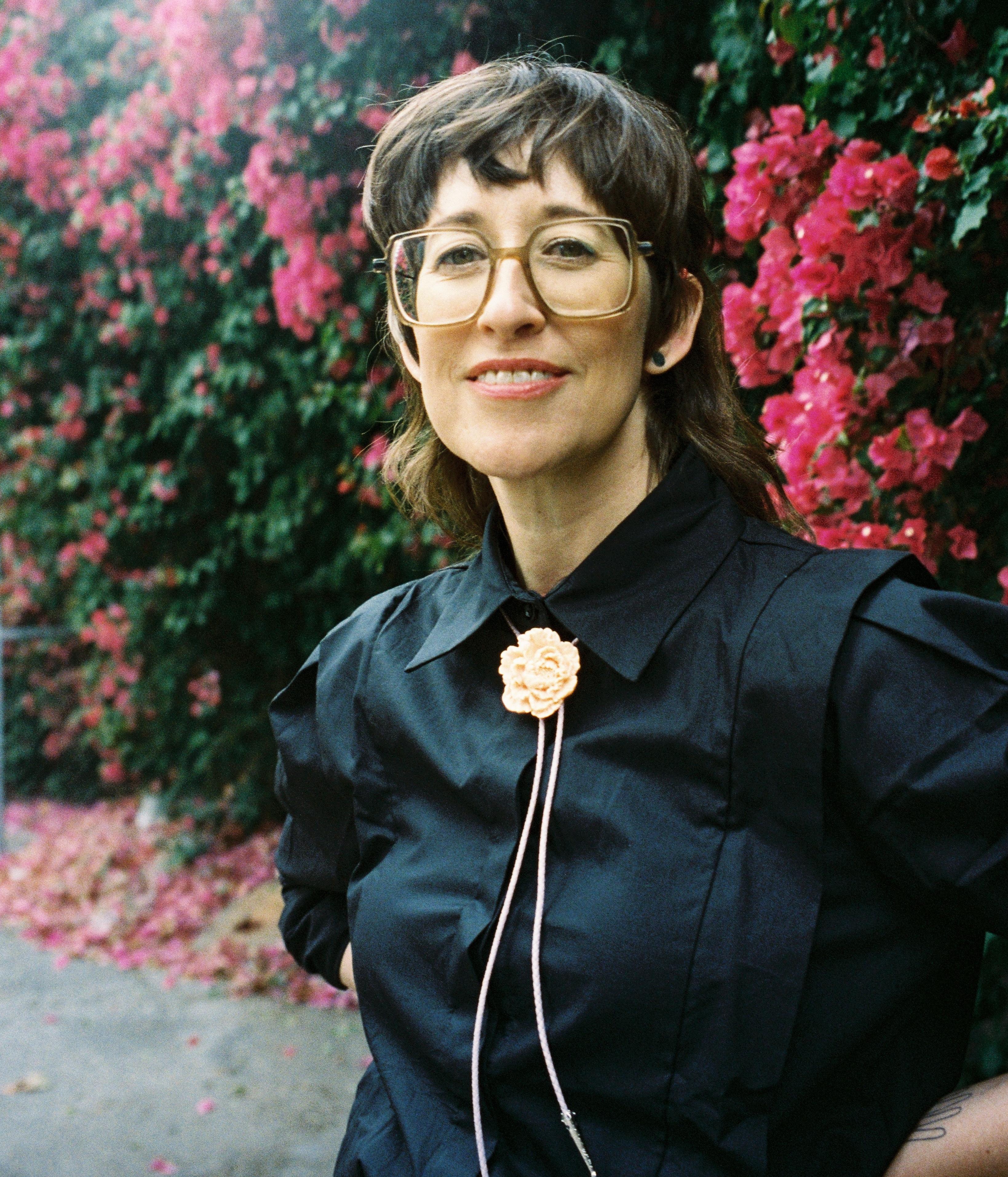 Alison is a smiling white woman with large brown glasses, long brown hair and windswept bangs wearing a black blouse and a peach flower bolo tie. She stands in front of a bougainvillea bush with bright pink flowers and her hands are on her hips.