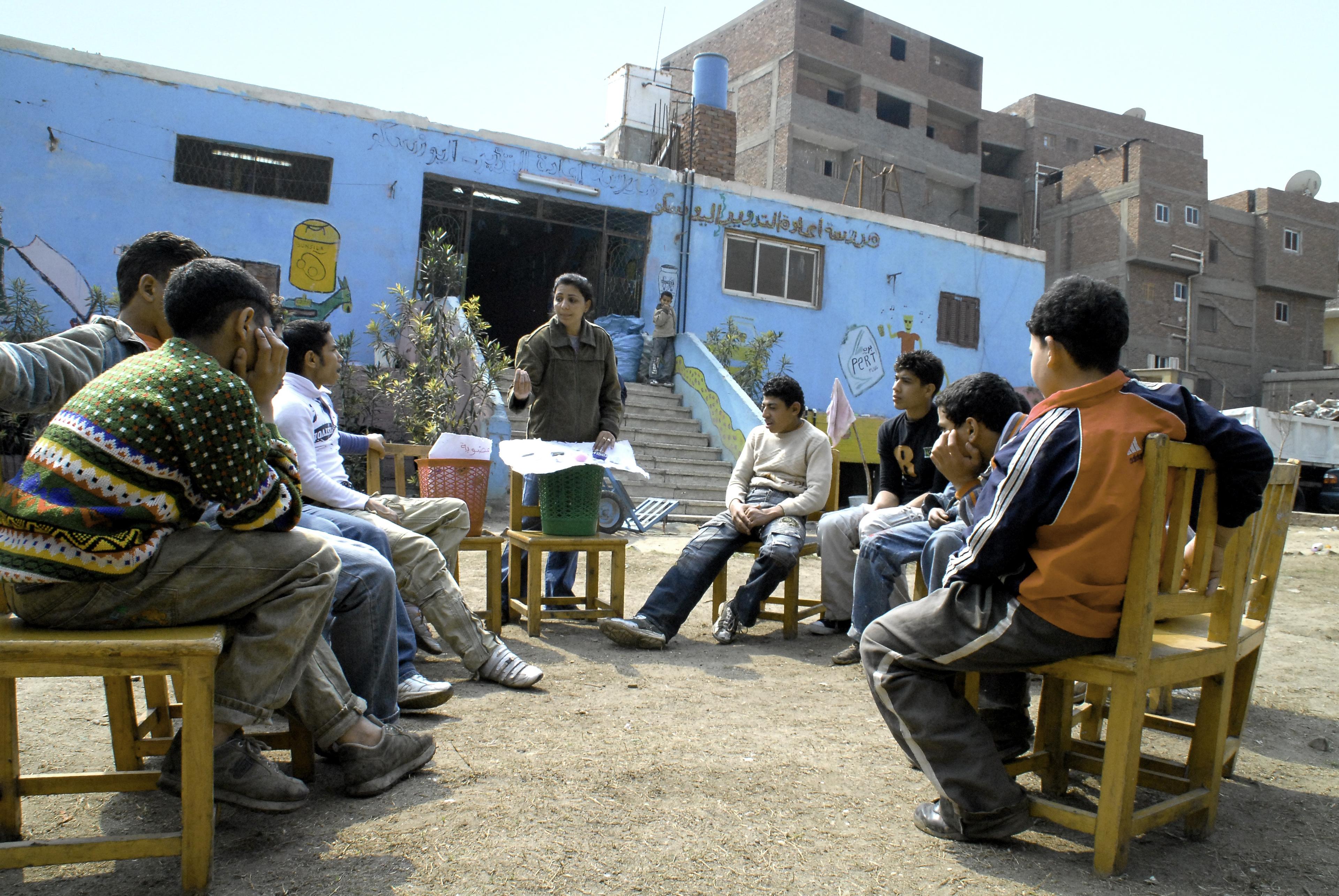 A group of students sit outside of school in a circle around trash cans while a teacher talks about recycling.