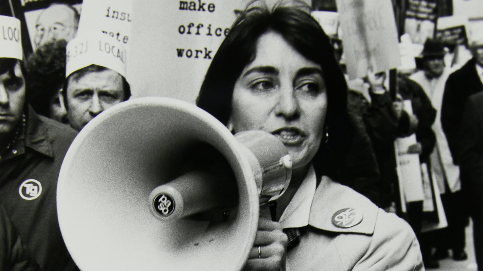 Still from 9to5: The Story of a Movement. Karen Nussbaum speaks into a megaphone at a Local 925 protest. Black and white image.